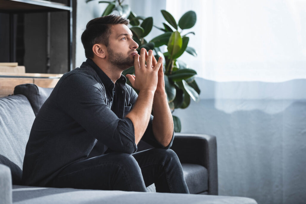 side view of handsome and pensive man sitting on sofa and looking up in apartment