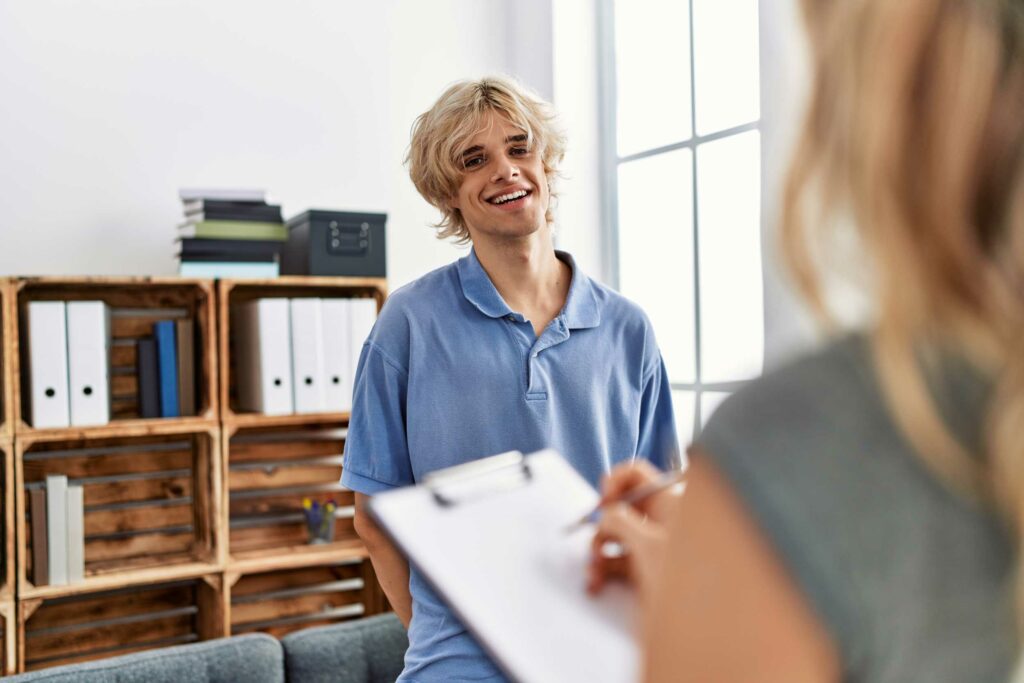 Young man with blonde hair smiling and holding a clipboard in an office, conversing with a person whose back is to the camera.