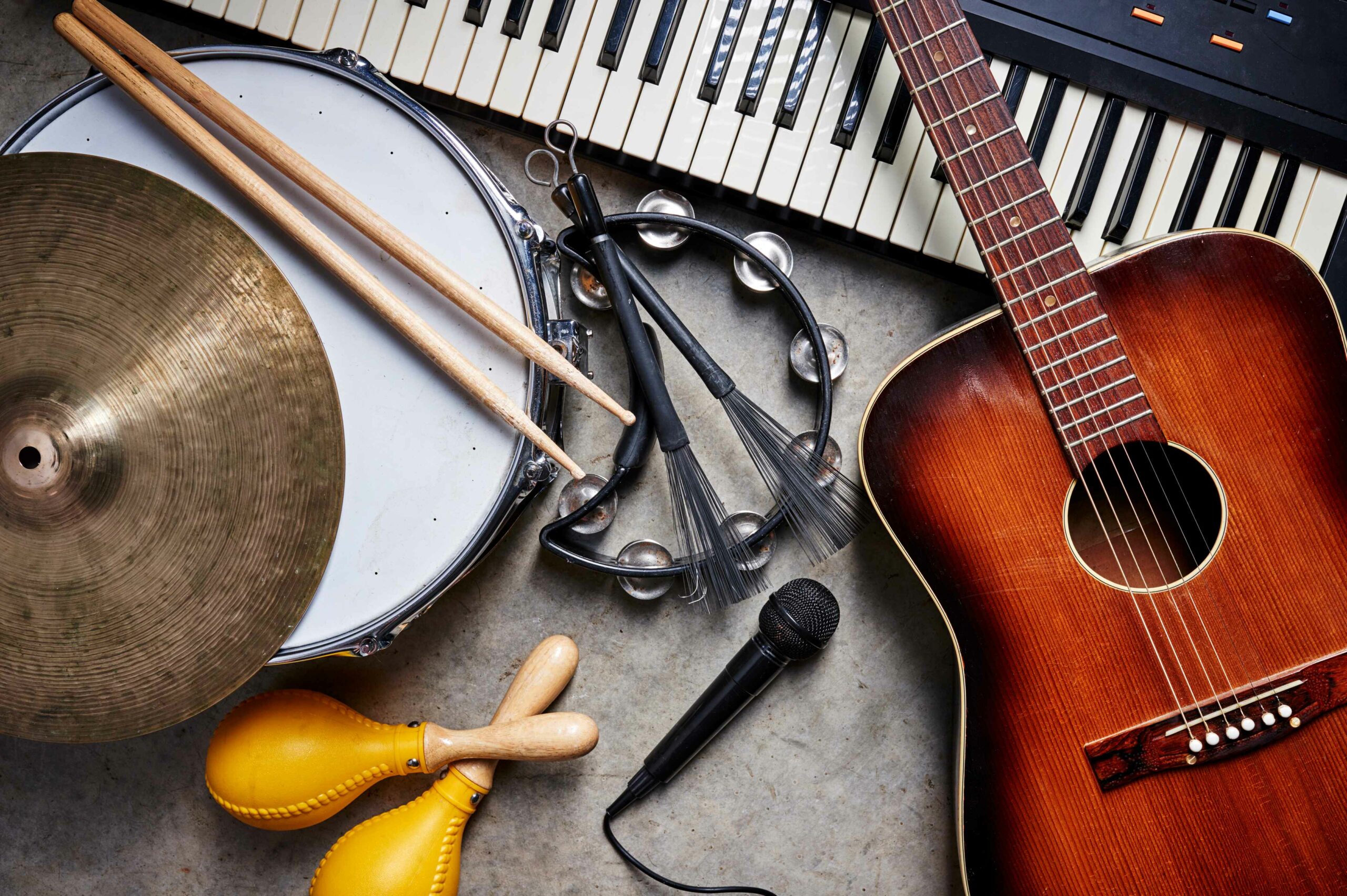 Instruments lay on cement floor including an acoustic guitar, a keyboard, a snare drum with drum sticks, a tambourine and other various percussion instruments